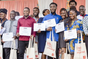 A group of people smiling while holding certificates, with bags at their feet, standing near curtains and a wall.