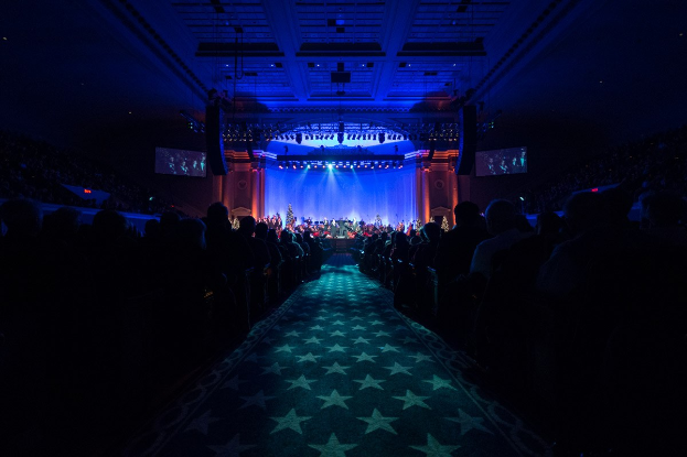 Ein großer Auditorium mit sitzenden Zuschauern, eine Bühne mit Musikern und Überkopfbeleuchtung und Fernseher, was auf einen Konzertsaal während der Nationalen Convention 2019 hinweist.