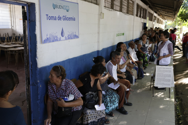 Menschen sitzen auf Bänken vor einem Gebäude, warten in der Schlange, um bei einer Wahlurne in Guadalajara, Mexiko, zu wählen, mit einer Tafel an der Wand, einer Tür links und Bäumen im Hintergrund.
