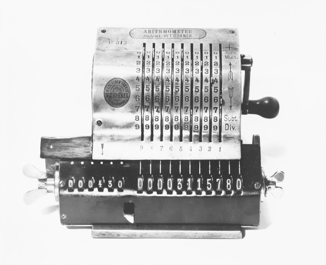 Altes Schwarz-Weiß-Foto eines klassischen elektronischen Kassenautomaten mit schwarzem Gehäuse, silbernem Deckel, großem Display, Tasten, Knöpfen, einem oberen Griff und einer seitlichen Tastatur auf einem weißen Hintergrund.