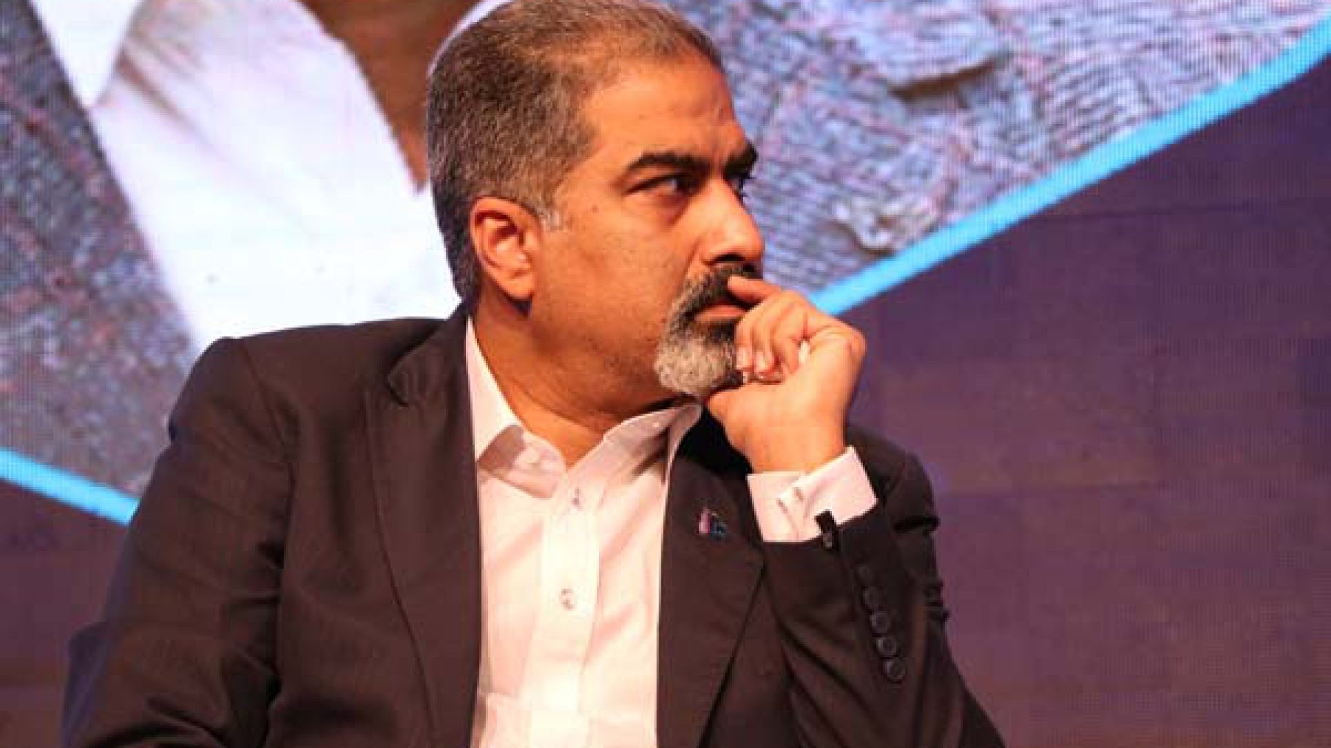 Ein Mann im Anzug und Krawatte sitzt auf einer Bühne, tief in Gedanken, vor einem Bildschirm.