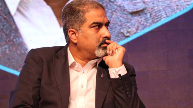 Ein Mann im Anzug und Krawatte sitzt auf einer Bühne, tief in Gedanken, vor einem Bildschirm.