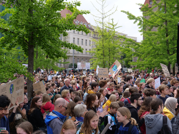 Eine große Menschenmenge protestiert vor einem Gebäude in Berlin und hält Schilder hoch, mit Bäumen, Fahrzeugen und einem Lautsprecher im Bild.