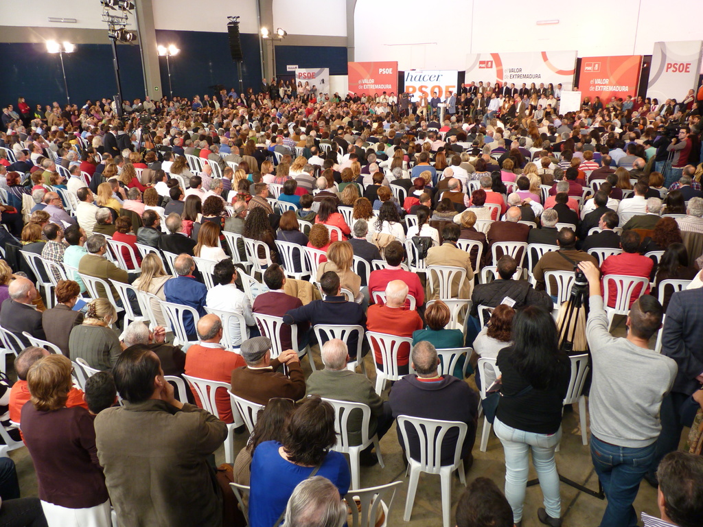 Ein volles Stadion mit vielen sitzenden und stehenden Menschen, eine B√ľhne mit zwei Rednern an einem Podium und mehreren Bannern und Lichtern in der N√§he.