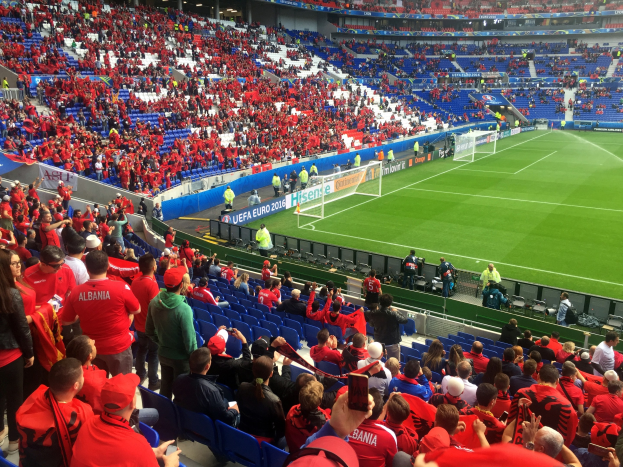 Ein volles Stadion mit Zuschauern, einige sitzen, andere stehen, mit Fahnen, einem Tor und Deckenbeleuchtung, während die Menge gespannt auf den Beginn des Fußballspiels wartet.