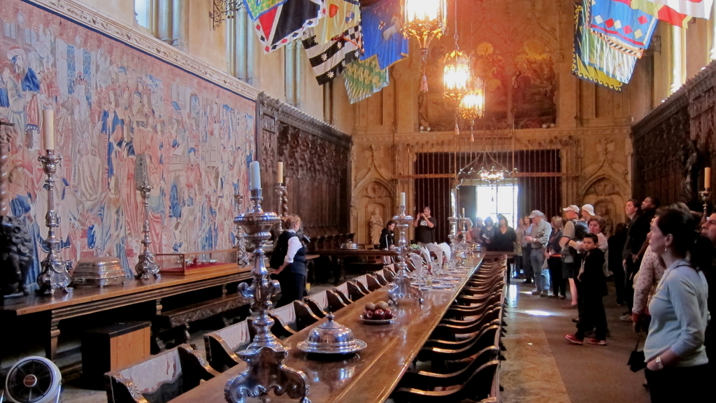 Ein großer Raum mit einem großen Esstisch, umgeben von vielen Stühlen, einer Kerze auf einem Ständer, einem Kronleuchter an der Decke und Plakaten und Flaggen im Hintergrund.