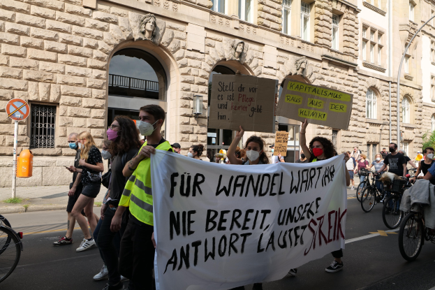 Eine Gruppe von Menschen marschiert bei einer Demonstration gegen die Friedensbewegung in Berlin, einige halten Schilder und andere fahren Fahrräder, im Hintergrund ist ein Gebäude mit Rundbogenfenstern, Säulen und Skulpturen zu sehen.