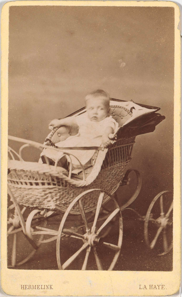 Ein altes Schwarz-Weiß-Foto eines Babys in einem hölzernen Kinderwagen mit vier Rädern, das ein weißes Strampler trägt und einen friedlichen Ausdruck hat, mit kursiver Schrift am unteren Rand.