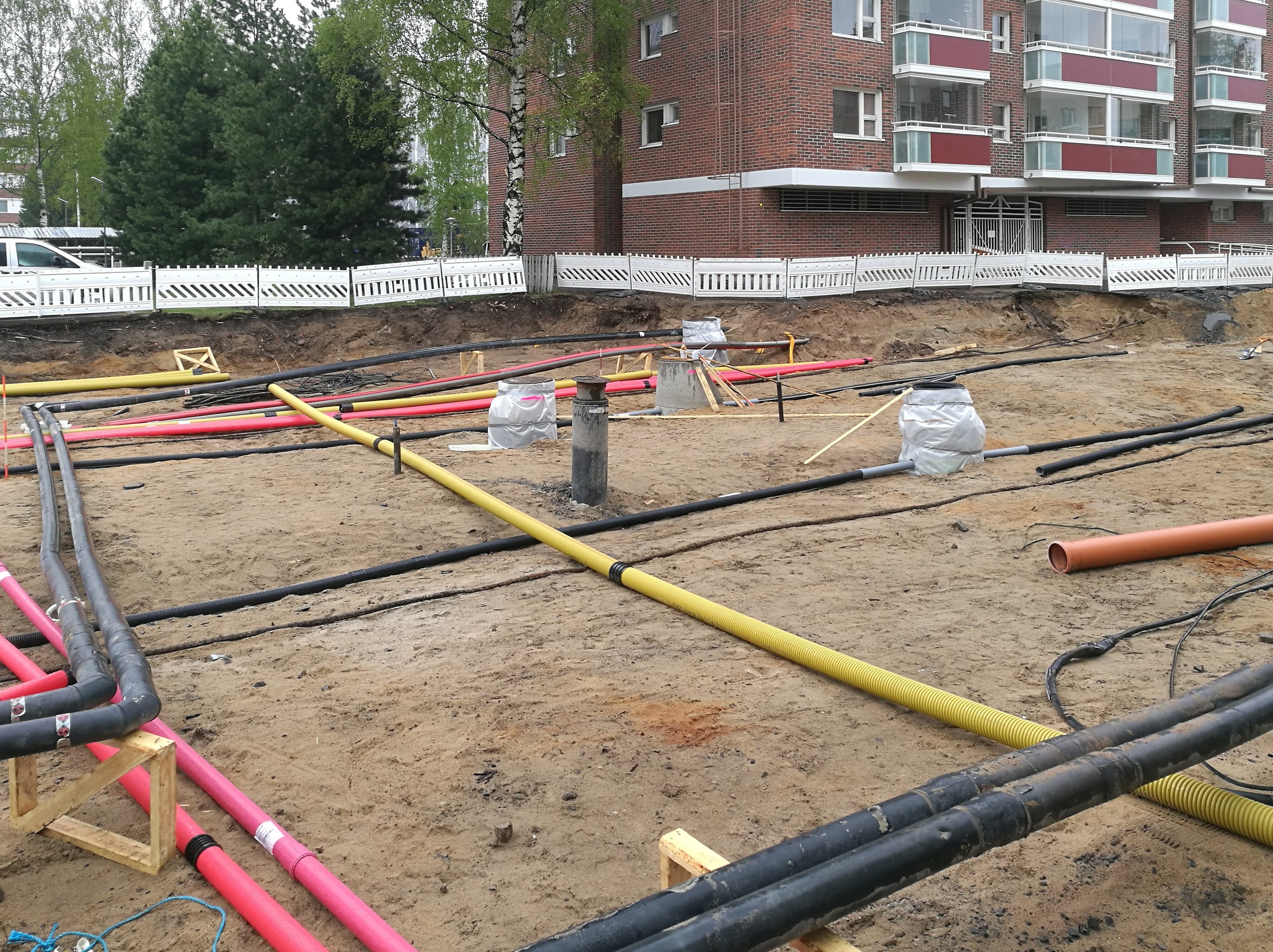 Großer Rohrstapel auf einer Baustelle mit verstreutem Material, umgeben von Gebäuden und Bäumen unter einem klaren blauen Himmel.