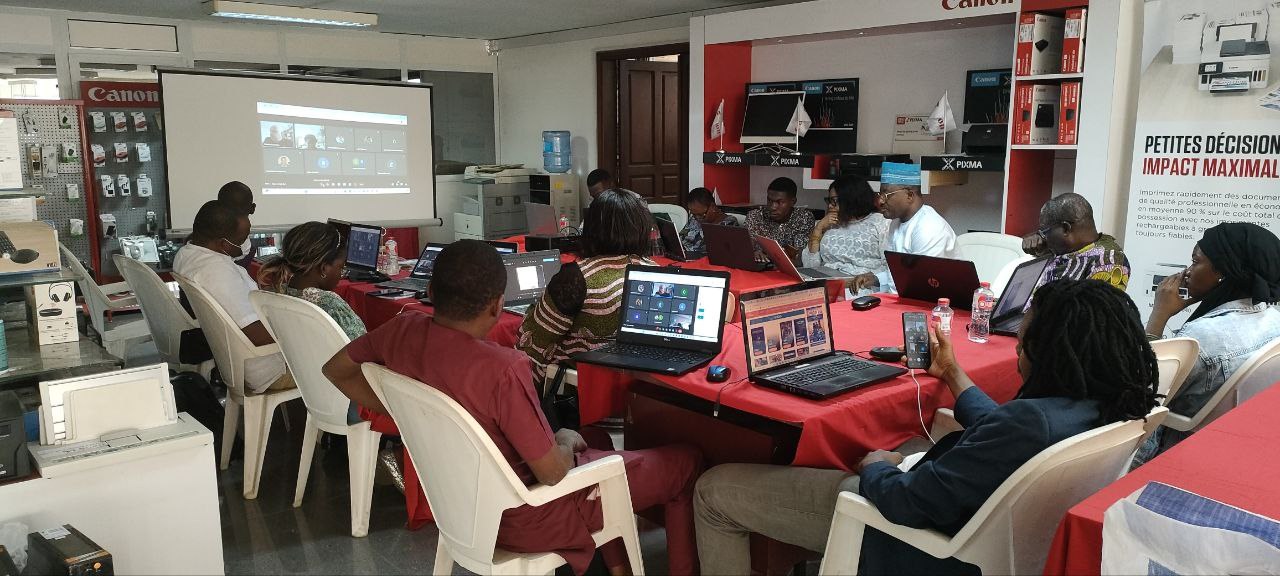 Gruppe von Menschen, die um einen Tisch mit Laptops und anderen Gegenständen während eines digitalen Marketing-Workshops sitzen, mit einem Banner, einem Bildschirm und Deckenleuchten im Raum.