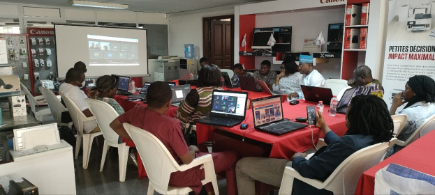 Gruppe von Menschen, die um einen Tisch mit Laptops und anderen Gegenständen während eines digitalen Marketing-Workshops sitzen, mit einem Banner, einem Bildschirm und Deckenleuchten im Raum.