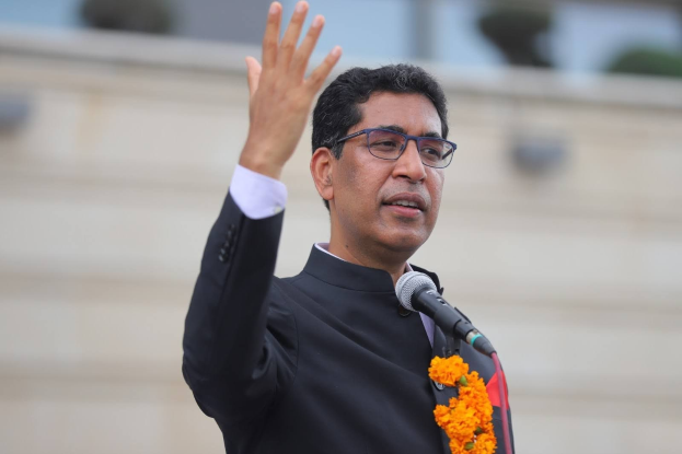 A man in a black suit and tie, wearing spectacles and a garland, speaks into a microphone during a speech.