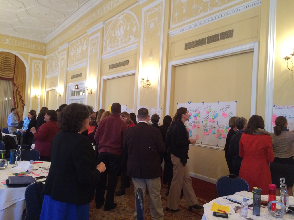 Eine Gruppe von Menschen untersucht Plakate und Diagramme, die an einer Wand in einem Raum zu sein scheinen, der ein Kunstmuseum zu sein scheint.