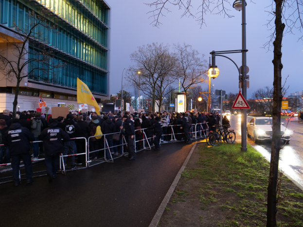 Large group of people protesting in front of a building in Berlin, holding banners, with barricades, bicycles, light poles, sign boards, trees, and grass visible.