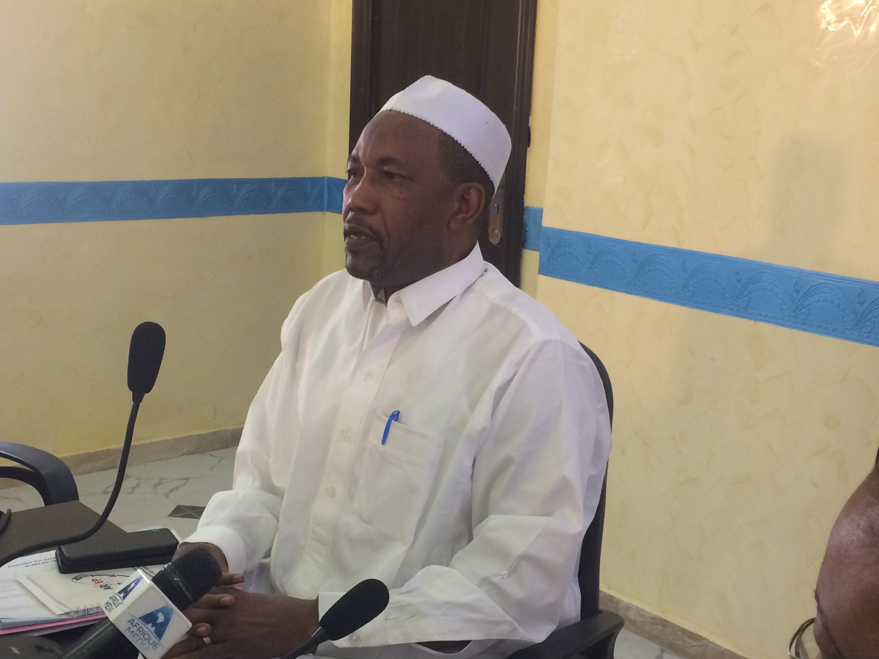 Ein Mann in einem weißen Kleid und Hut sitzt an einem Tisch mit Mikrofonen und spricht, während Papiere und Gegenstände auf dem Tisch sichtbar sind; rechts von ihm sitzt eine Person mit Brille, im Hintergrund eine Tür und eine Wand.