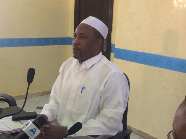 Ein Mann in einem weißen Kleid und Hut sitzt an einem Tisch mit Mikrofonen und spricht, während Papiere und Gegenstände auf dem Tisch sichtbar sind; rechts von ihm sitzt eine Person mit Brille, im Hintergrund eine Tür und eine Wand.