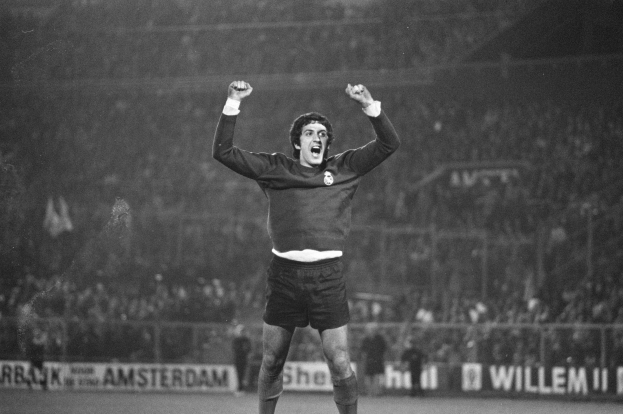 Schwarzes und weißes Foto eines Mannes, der auf einem Fußballfeld feiert, die Arme erhoben, umgeben von einer Menge, einem Zaun und Bannern mit Text.