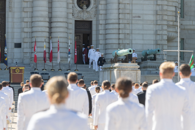 Gruppe von Menschen in weißen Marineuniformen vor einem Gebäude mit Säulen und Treppen, mit Fahnen, einem Podium und Kanonen im Hintergrund.