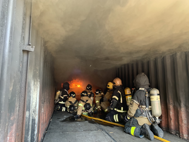 Feuerwehrleute mit Helmen und Sauerstoffflaschen arbeiten in einem raucherfüllten Tunnel, einer hält ein Rohr, mit sichtbarem Feuer und einer Wand links.