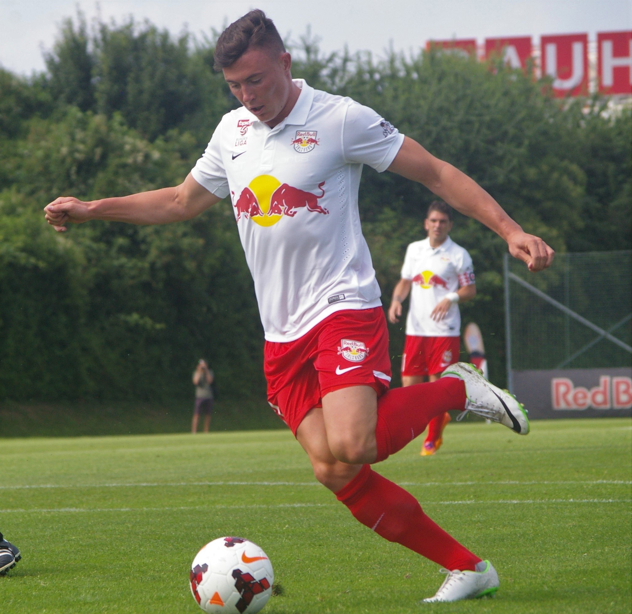 A professional soccer player kicks a ball on a grassy field with "RB Leipzig" visible in the background under a clear blue sky.