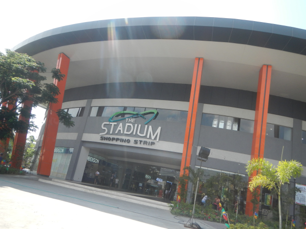 Large glass-walled shopping mall with a prominent name sign, surrounded by greenery and people walking outside under a visible sky.