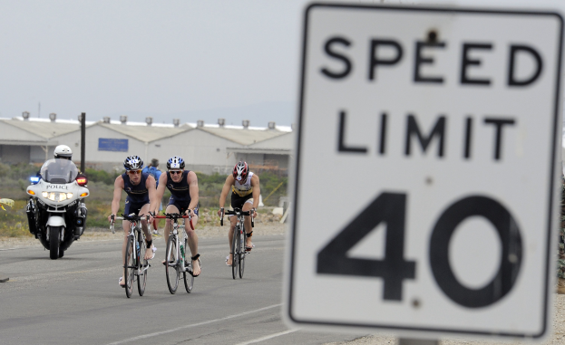 Gruppe von Radfahrern in Helmen, die auf einer Straße mit einem Tempolimitschild fahren, Gebäude und Bäume im Hintergrund unter einem klaren blauen Himmel.