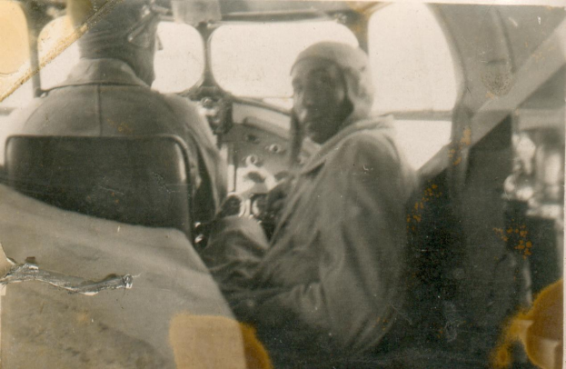 Schwarzes und weißes Foto von zwei Männern im Cockpit eines Flugzeugs; einer sitzt in Pilotenuniform, der andere steht neben ihm, beide mit ernsten Gesichtern.
