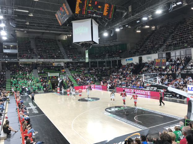 Basketballspiel in einer großen Arena mit Publikum, Anzeigetafeln mit "Asvel vs Galatasaray" und Deckenbeleuchtung und Bildschirme.