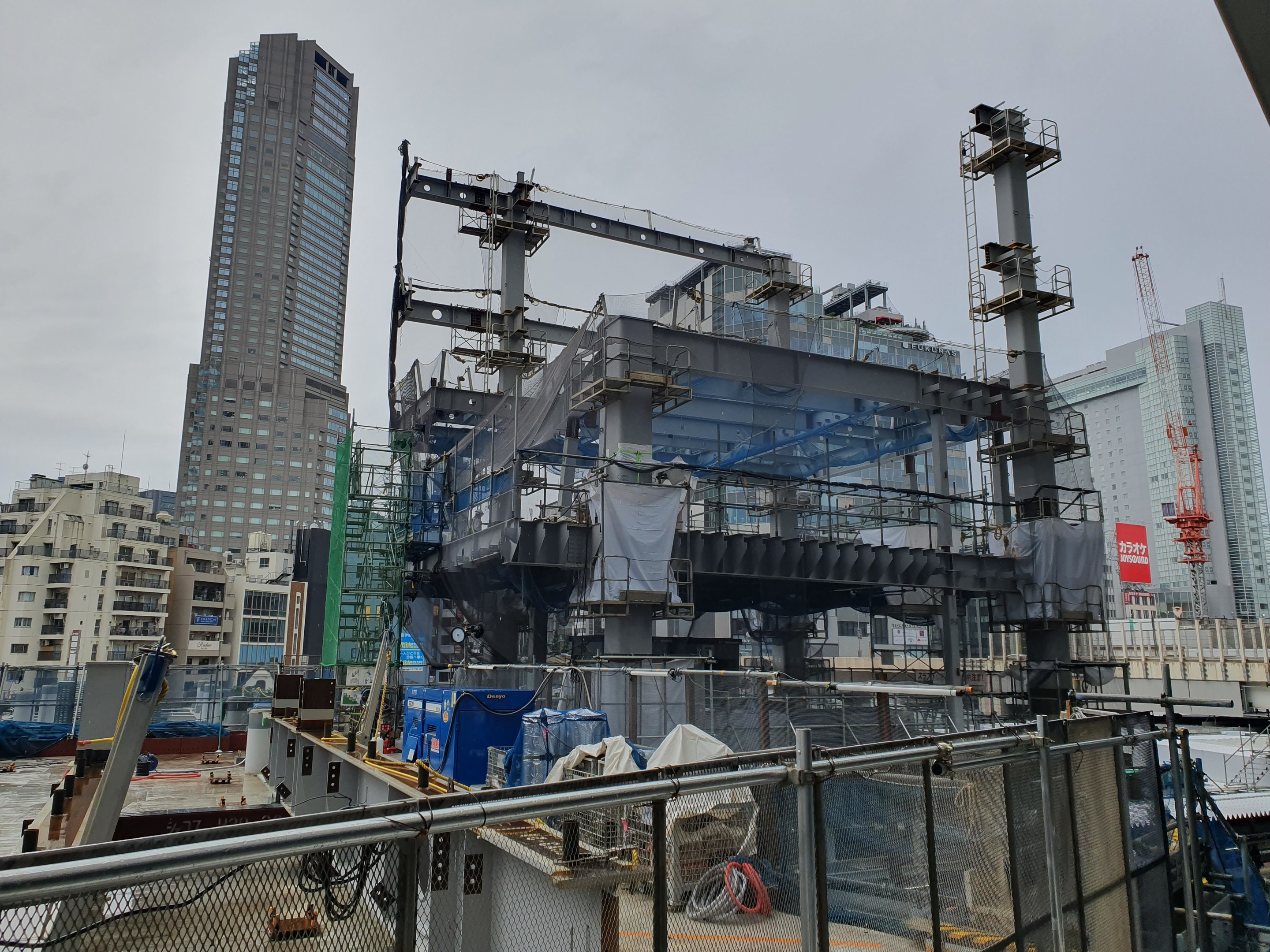 Großbaustelle für ein neues Kraftwerk in einer Stadtumgebung, umgeben von einem Metallzaun, mit sichtbaren Gebäuden, Fahrzeugen, Polen, Drähten, einer Fahne und einem bewölkten Himmel.