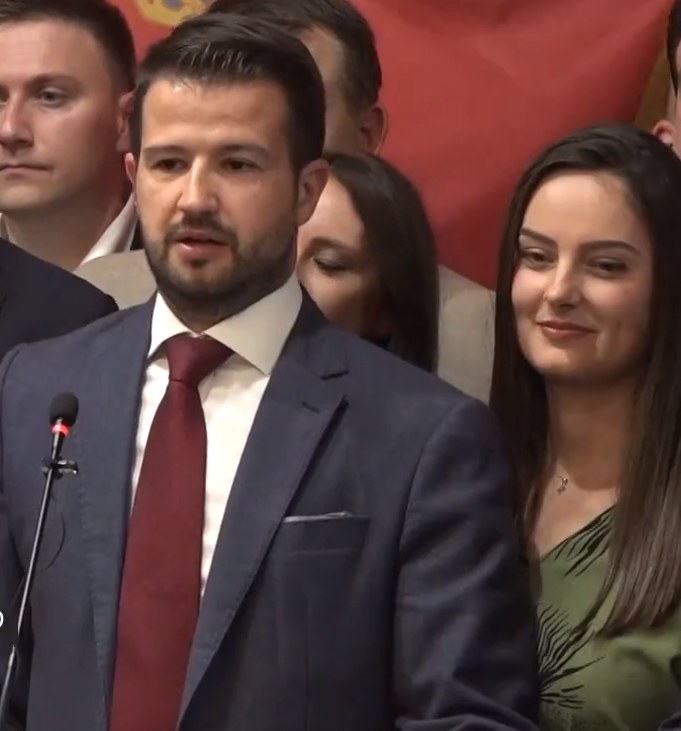 Ein Mann im Anzug steht an einem Rednerpult und spricht in ein Mikrofon, mit einem Banner im Hintergrund und einer sichtbaren Menge, wahrscheinlich wegen eines sozialen Medienverbots.