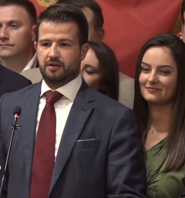 Ein Mann im Anzug steht an einem Rednerpult und spricht in ein Mikrofon, mit einem Banner im Hintergrund und einer sichtbaren Menge, wahrscheinlich wegen eines sozialen Medienverbots.