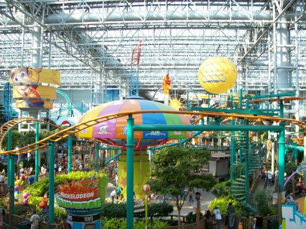 Ein großes Indoor-Wasserpark mit einer Achterbahn, umgeben von Menschen, Bäumen, Pflanzen und Deckenleuchten, die eine lebendige Atmosphäre schaffen.