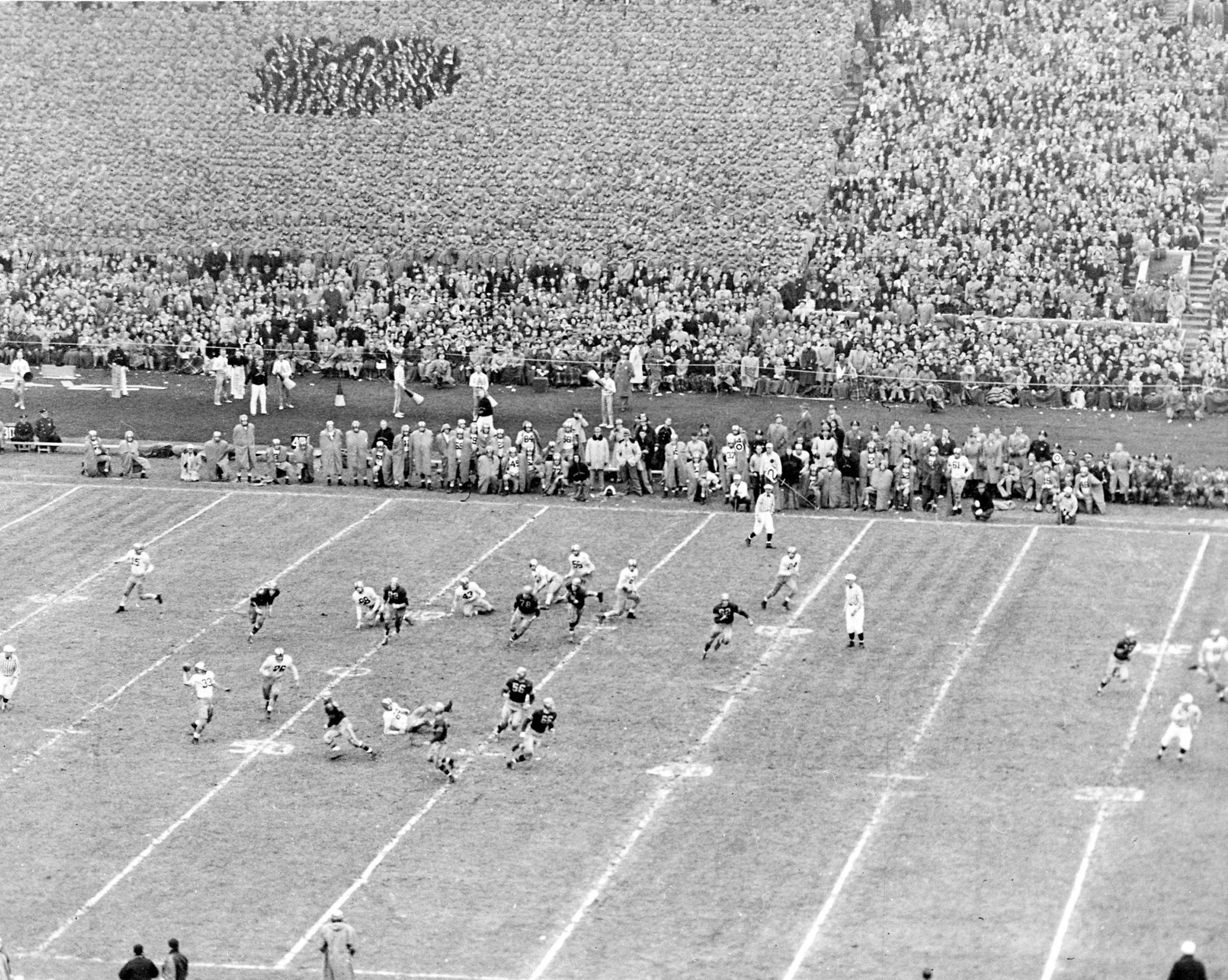 Schwarzes und weißes Foto eines Fußballspiels im Gange, mit Spielern auf dem Feld und einer Zuschauermenge im Hintergrund.
