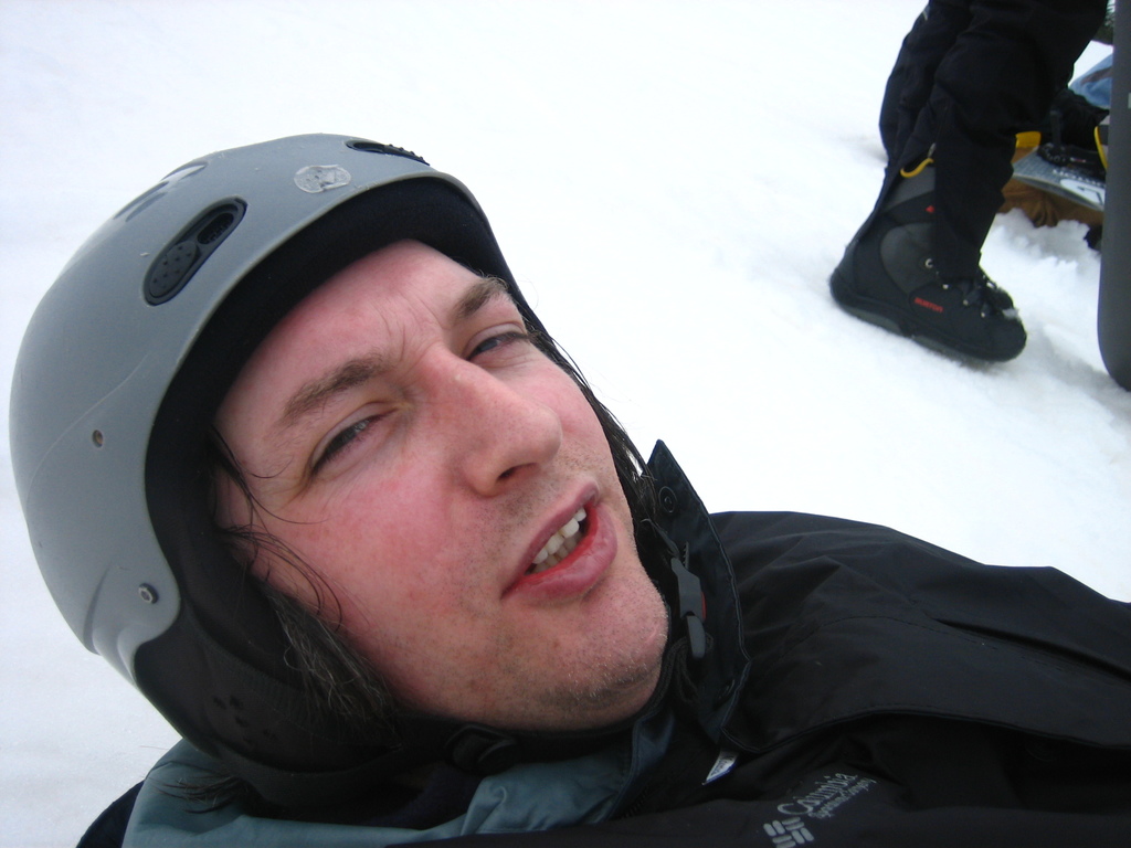 Ein Mann in einem Pullover und Helm steht im Schnee, mit den Beinen und Skiern einer anderen Person in der oberen rechten Ecke.