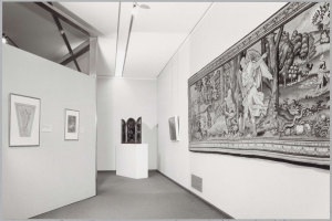 Black and white photo of a museum room with a large tapestry, framed art on walls, pedestal objects, ceiling lights, and "Museum of Fine Arts, New York City" text at the bottom.