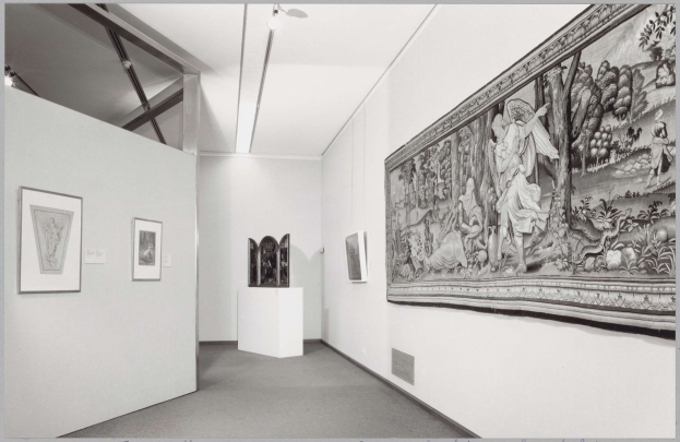 Black and white photo of a museum room with a large tapestry, framed art on walls, pedestal objects, ceiling lights, and "Museum of Fine Arts, New York City" text at the bottom.