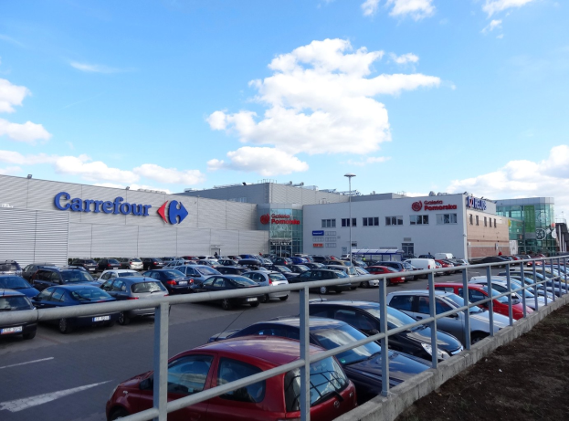 Ein großes Autohaus mit zahlreichen parkenden Fahrzeugen, umgeben von Gebäuden, Straßenlaternen, Schildern, Motorrädern auf der Straße, Gittern und einem bewölkten Himmel im Hintergrund.