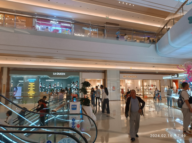 Interior of a shopping mall with people walking, escalators, illuminated ceiling lights, decorative plants, informational boards, storefronts, and shoppers carrying bags, with a watermark noting its recent reopening post-coronavirus pandemic.