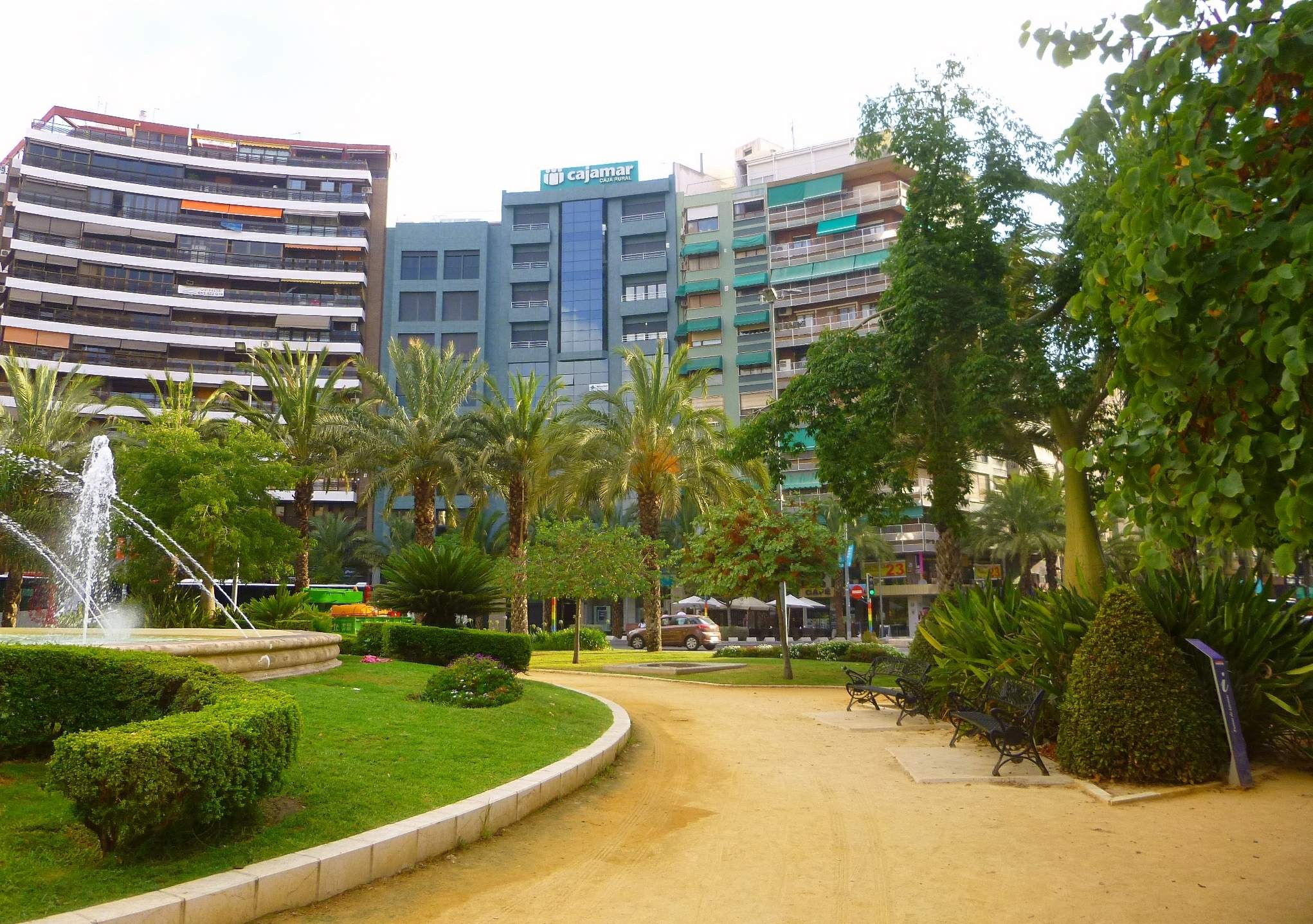 Ein Park mit einem zentralen Brunnen, umgeben von Grünflächen, Bänken, Laternen und Fahrzeugen auf einer Straße, mit hohen Gebäuden und einem klaren Himmel im Hintergrund.