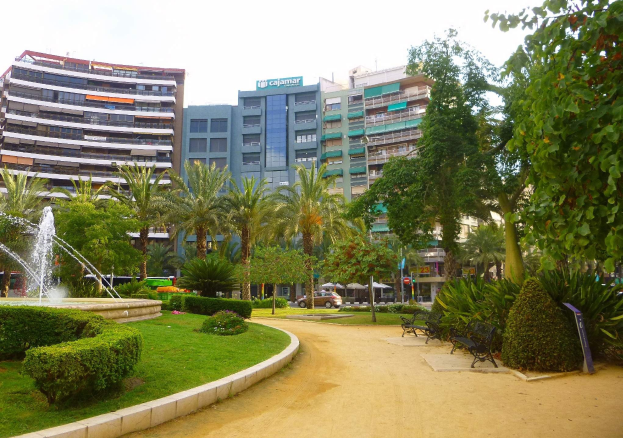 Ein Park mit einem zentralen Brunnen, umgeben von Grünflächen, Bänken, Laternen und Fahrzeugen auf einer Straße, mit hohen Gebäuden und einem klaren Himmel im Hintergrund.