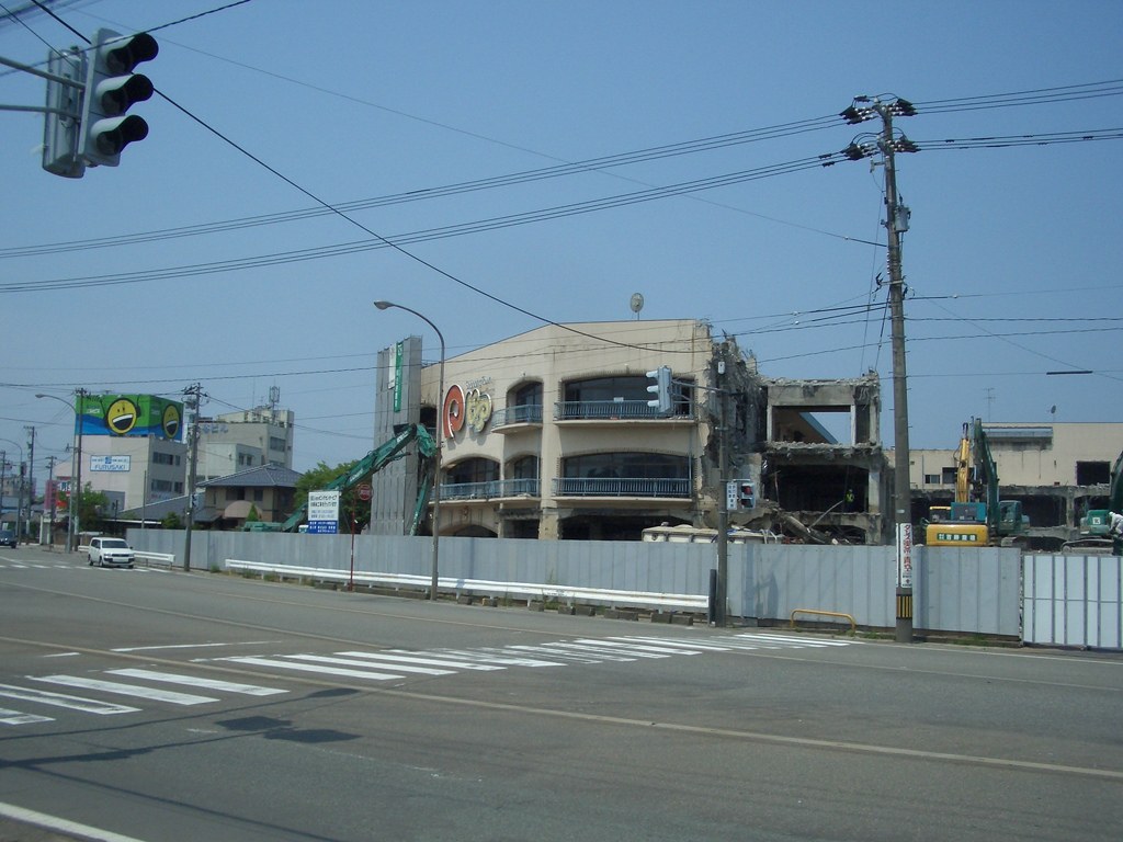 Ein abgerissenes Gebäude mit einem Metallzaun, einem Schild, Fahrzeugen, Mülltonnen, Pfählen, einem Banner an einer Wand, Bäumen und einem bewölkten Himmel.