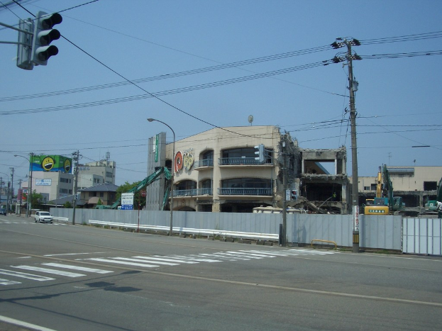 Ein abgerissenes Gebäude mit einem Metallzaun, einem Schild, Fahrzeugen, Mülltonnen, Pfählen, einem Banner an einer Wand, Bäumen und einem bewölkten Himmel.