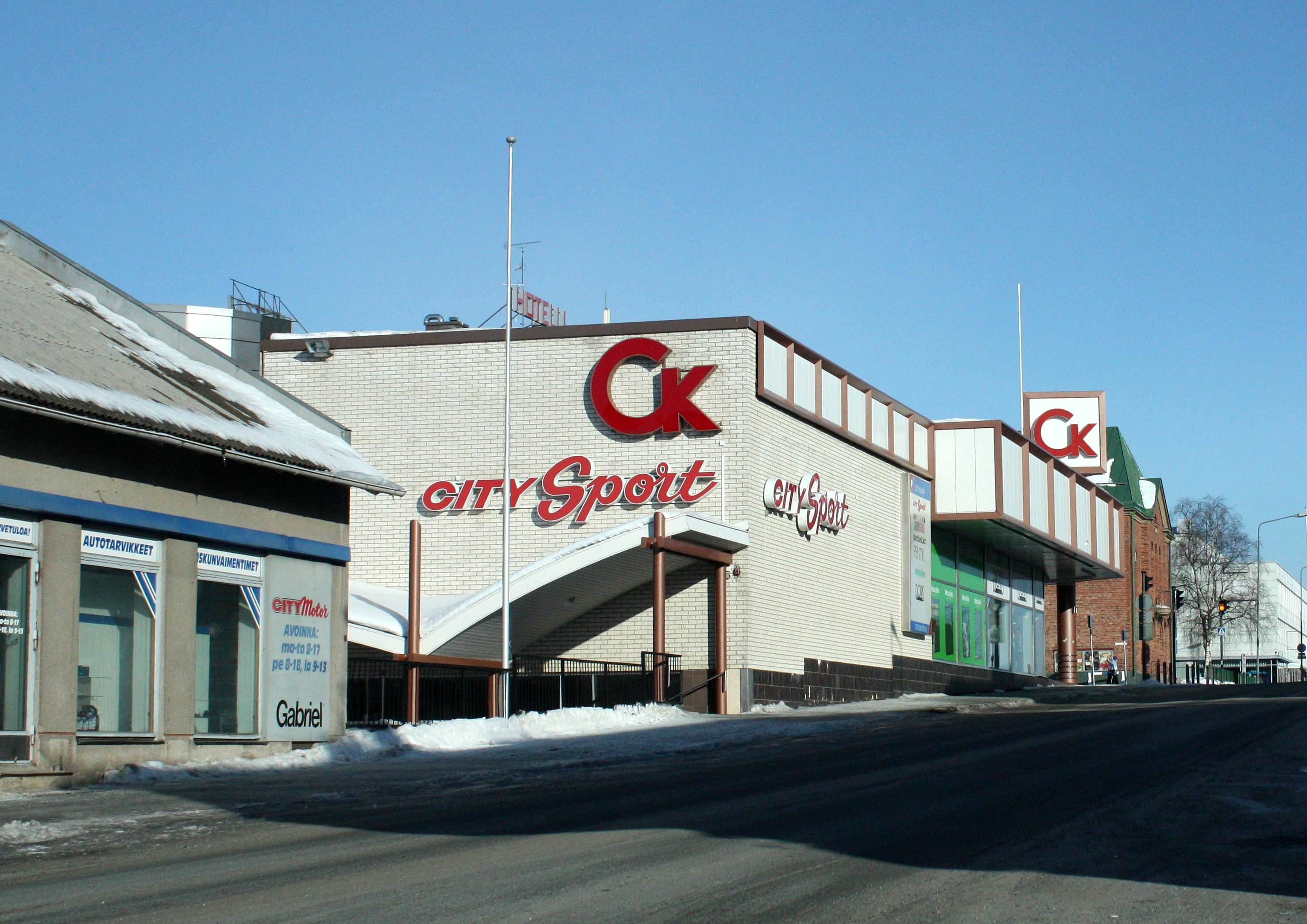 Ein Stadt-Sportgeschäft an einer verschneiten Straßenecke, umgeben von Gebäuden, Straßenmöbeln und Bäumen, mit dem Himmel im Hintergrund.