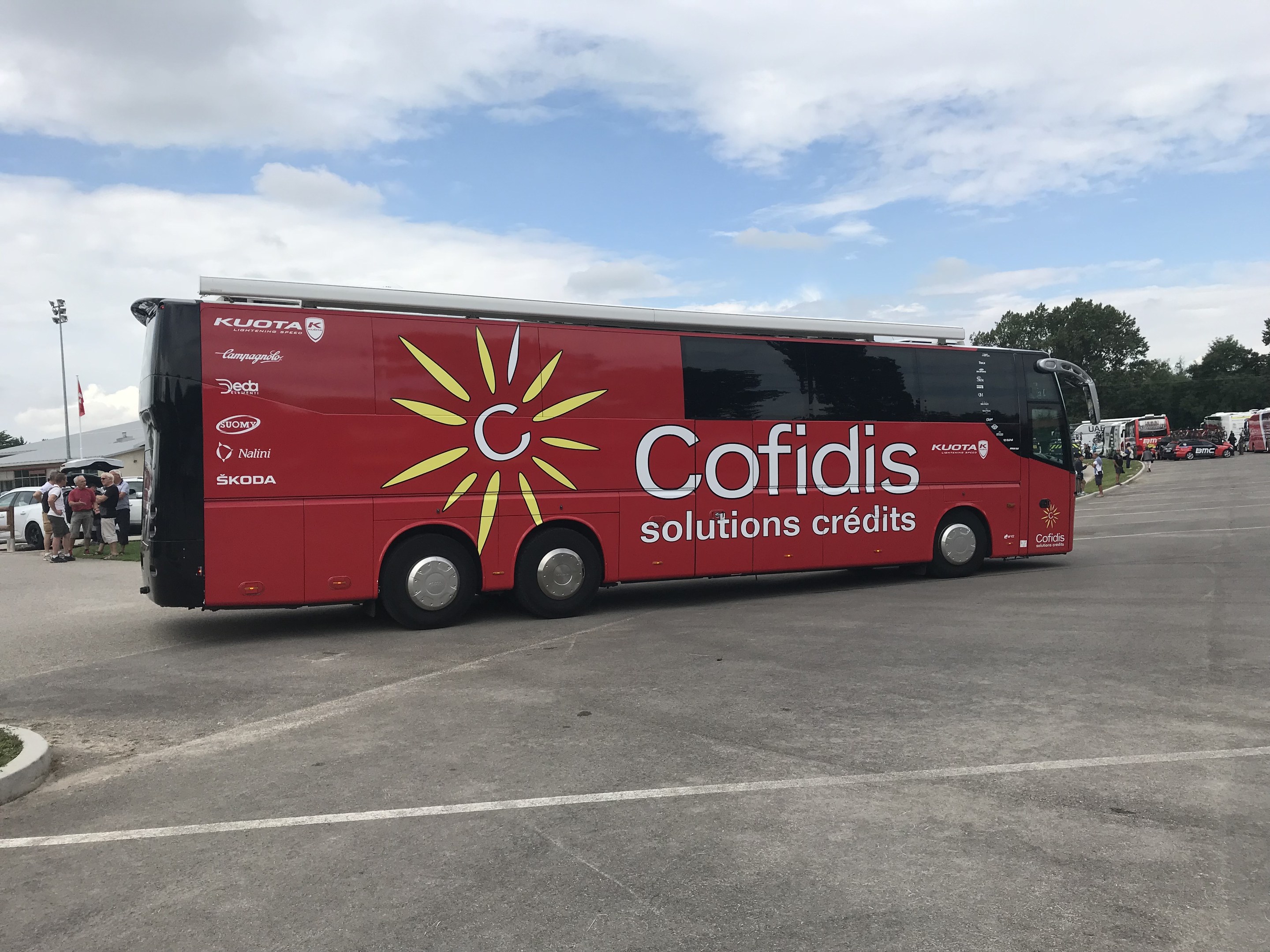 Roter Bus auf einem Parkplatz mit Menschen in der Nähe, Fahrzeugen auf der Straße, einem Straßenschild, einem "Cofidis Solutions Credit"-Schild, Bäumen und einer bewölkten Himmel.