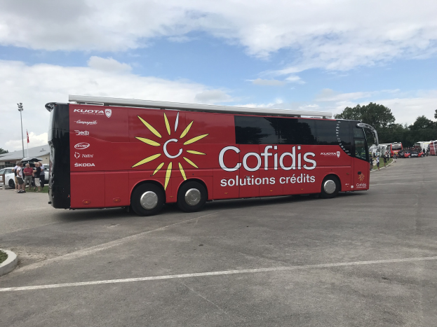 Roter Bus auf einem Parkplatz mit Menschen in der Nähe, Fahrzeugen auf der Straße, einem Straßenschild, einem "Cofidis Solutions Credit"-Schild, Bäumen und einer bewölkten Himmel.