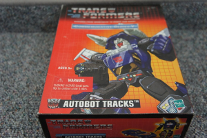 Ein roter, oranger und blauer Autobot Tracks Roboter-Spielzeugkasten mit einem Roboter oben drauf und Text, vor einem unscharfen schwarzen Hintergrund.