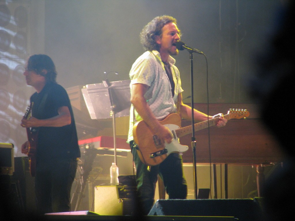 Live music performance on stage with a man in a white shirt singing into a microphone and playing guitar, accompanied by another man in a black shirt playing guitar, with piano, cables, speakers, and stage lights in the background.