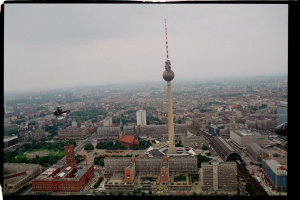 Luftaufnahme von Berlin, Deutschland, mit dem Fernsehturm in der Mitte, umgeben von Gebäuden, Bäumen, Straßen, Fahrzeugen und einem Hubschrauber unter einem klaren blauen Himmel.