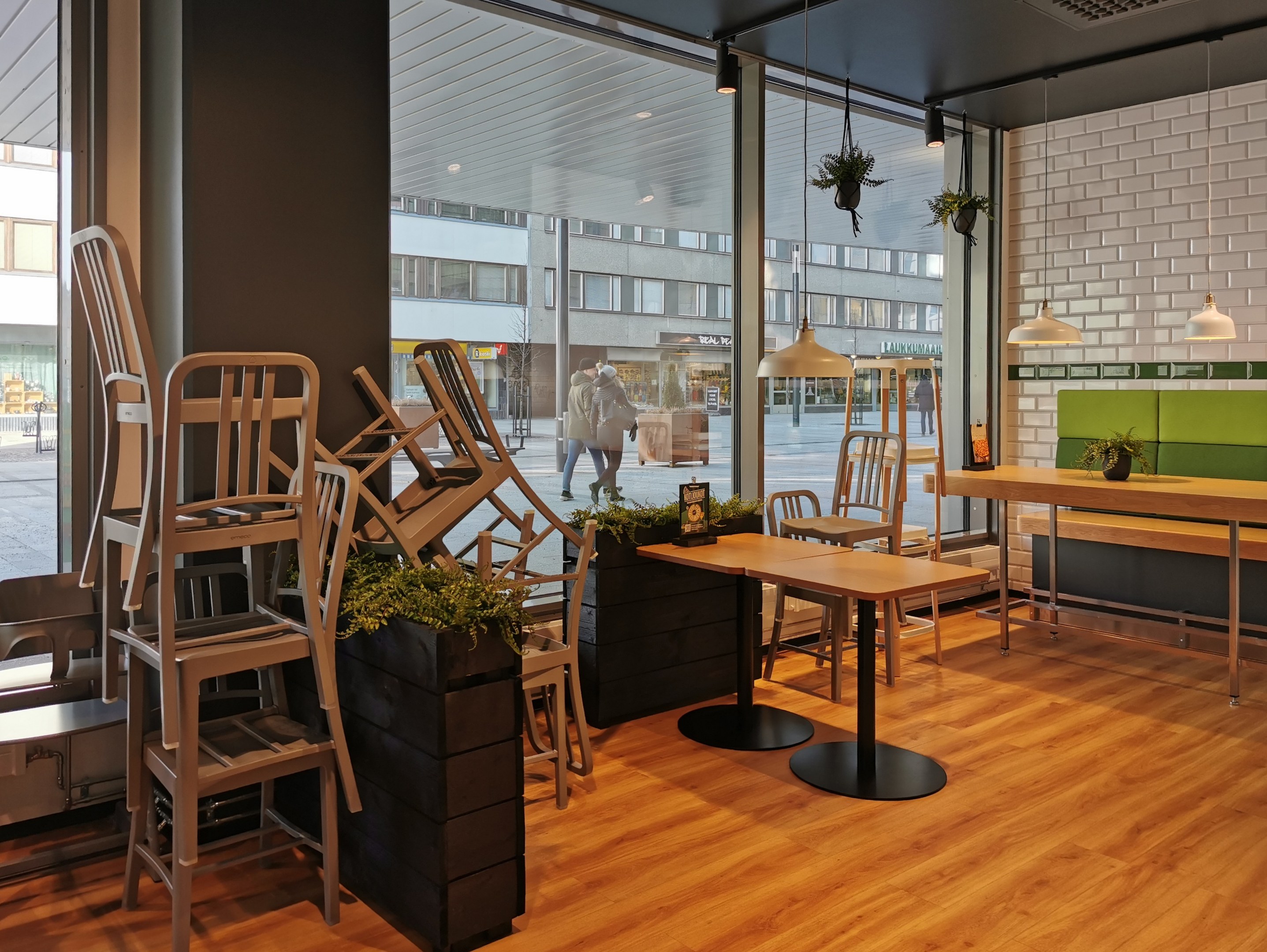 Ein Restaurant mit hölzernen Tischen und Stühlen drinnen, durch ein großes Fenster mit einer Berliner Straßenszene mit Passanten, Gebäuden und urbanen Elementen.