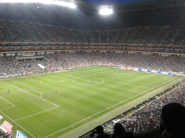 Ein hell erleuchtetes Fußballstadion voller jubelnder Fans, die ein Spiel verfolgen, mit Werbetafeln und elektrisierender Atmosphäre.